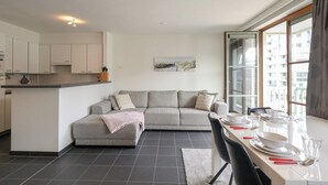 A modern living room with a grey sofa, a dining table set with plates and glasses, and a kitchen area with white cabinets.
