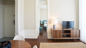 A hotel room with a wooden TV stand, a television, a small table, and a chair. There is a door leading to another room and a hallway visible in the background.