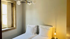 A single bed with white bedding, a bedside table with a lamp, and a window with a view of the outside. - Cozy City Family Retreat (Oporto)