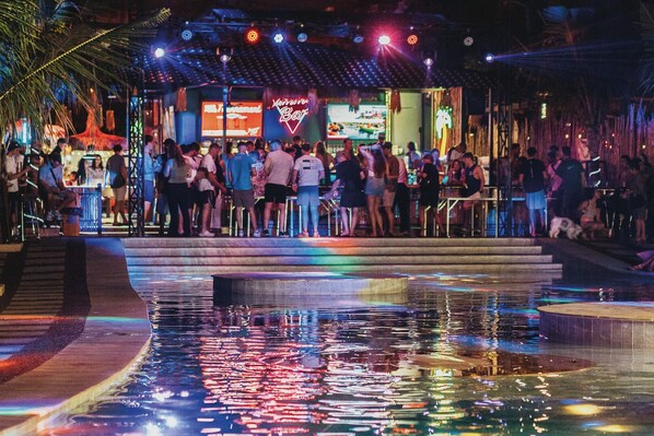 Ein belebter Außenbereich einer Bar mit Pool, der Lichter spiegelt, umgeben von Palmen und einem Neonzeichen.