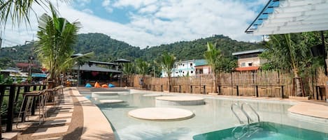 A swimming pool with circular platforms, surrounded by palm trees and a bamboo fence.
