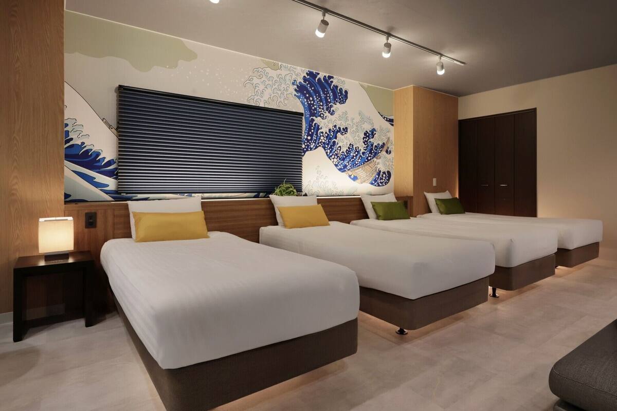 Une chambre d’hôtel avec deux lits, une grande fenêtre avec stores, une fresque murale et une table de chevet avec une lampe.