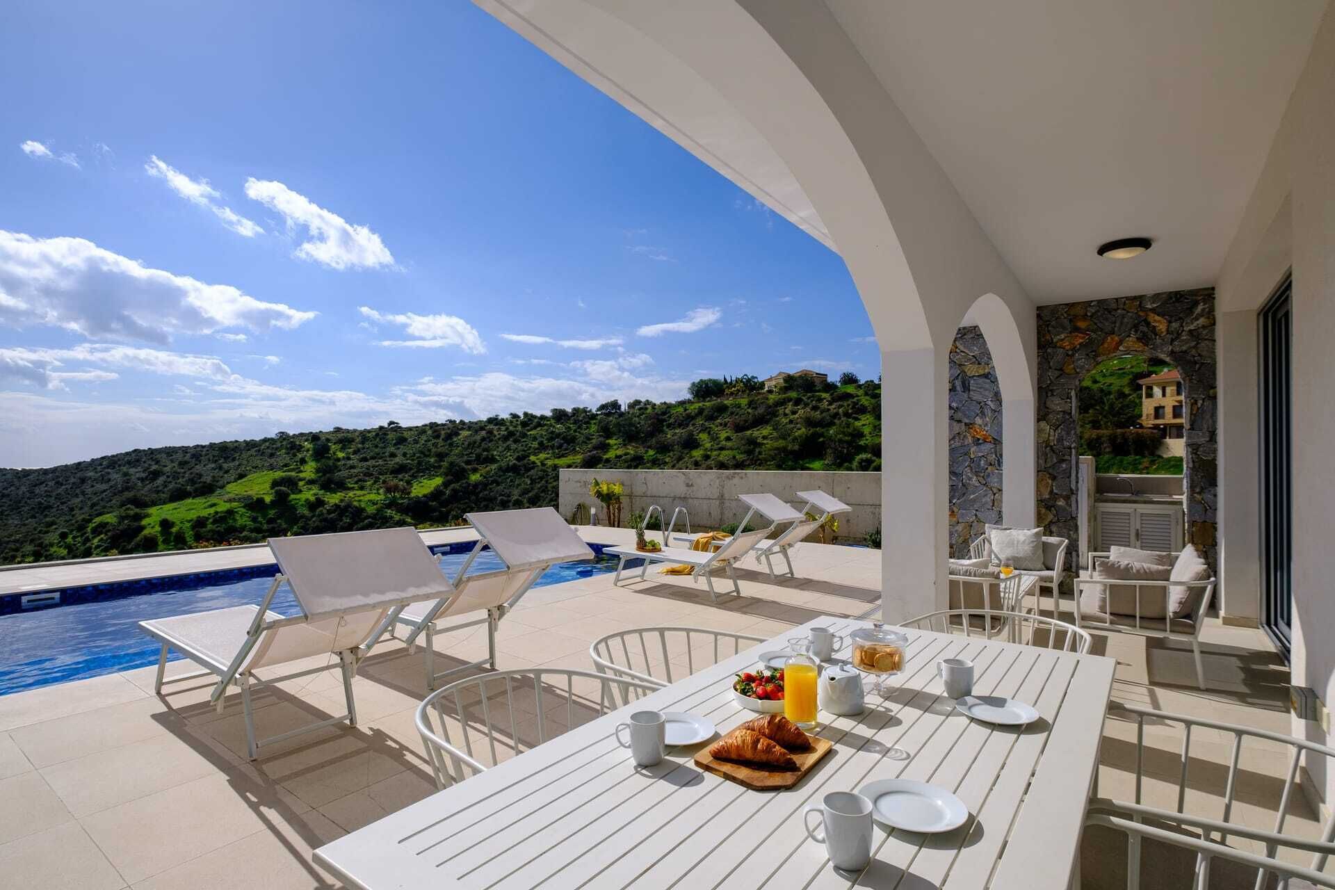 Une terrasse avec une piscine, des chaises longues, une table à manger où le petit-déjeuner est servi, et une vue panoramique sur les collines.