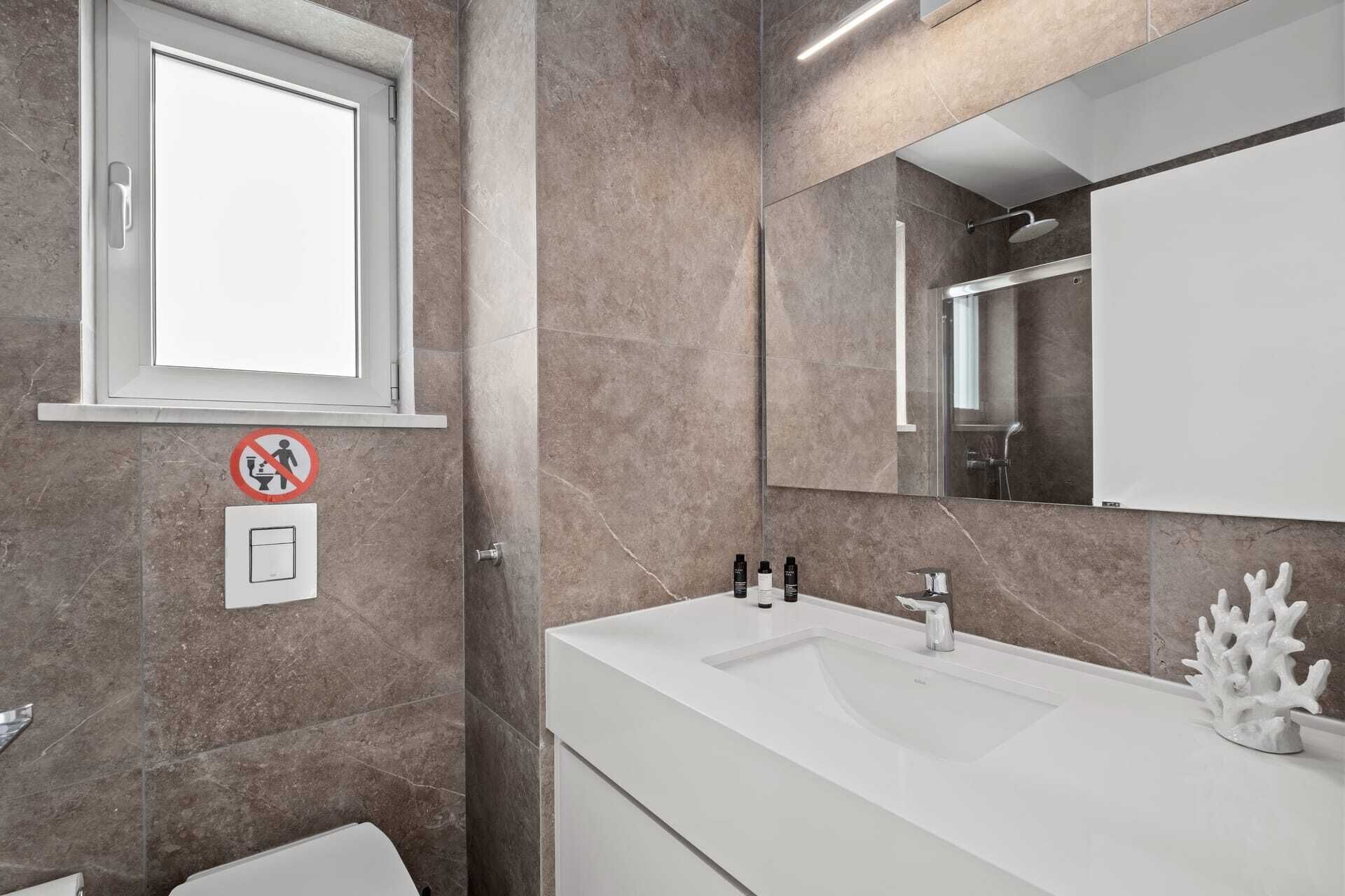 Une salle de bain moderne avec un grand miroir, un lavabo blanc et une douche. On trouve des articles de toilette sur le lavabo et un panneau «interdiction de fumer» sur le mur.