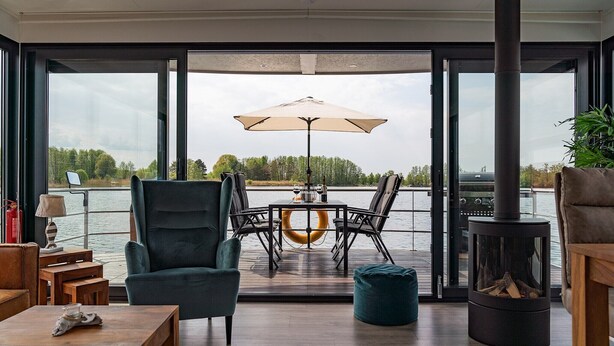 A modern living room with a large window, a dining table with chairs, a sofa, a small table, and a fireplace. - Havel Houseboat Serenity (Zehdenick)
