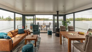 A spacious indoor area with a large window overlooking a body of water, furnished with a sofa, chairs, a table, and a fireplace. - Havel Houseboat Serenity (Zehdenick)