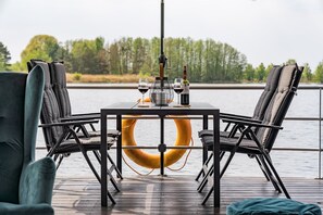 Een tafel met twee stoelen, een reddingsboei en twee glazen wijn op een terras met uitzicht op een meer en bomen.