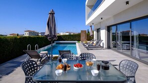 A modern outdoor pool area with lounge chairs, a dining table with breakfast items, and a clear view of the pool. - Soleil Luxury Villa by Nomads (Pegeia)