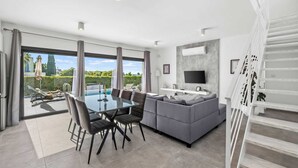 A modern living space with a dining area, a sofa, and a TV. There is a pool visible through the glass doors. - Soleil Luxury Villa by Nomads (Pegeia)