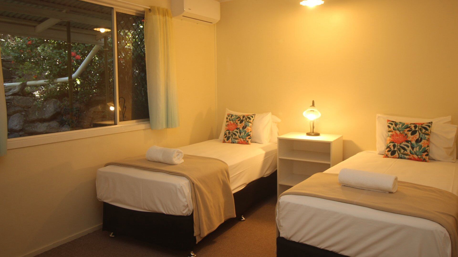 A room with two beds, each with a pillow and a bedside table with a lamp. There is a window with a curtain and an air conditioning unit above it.