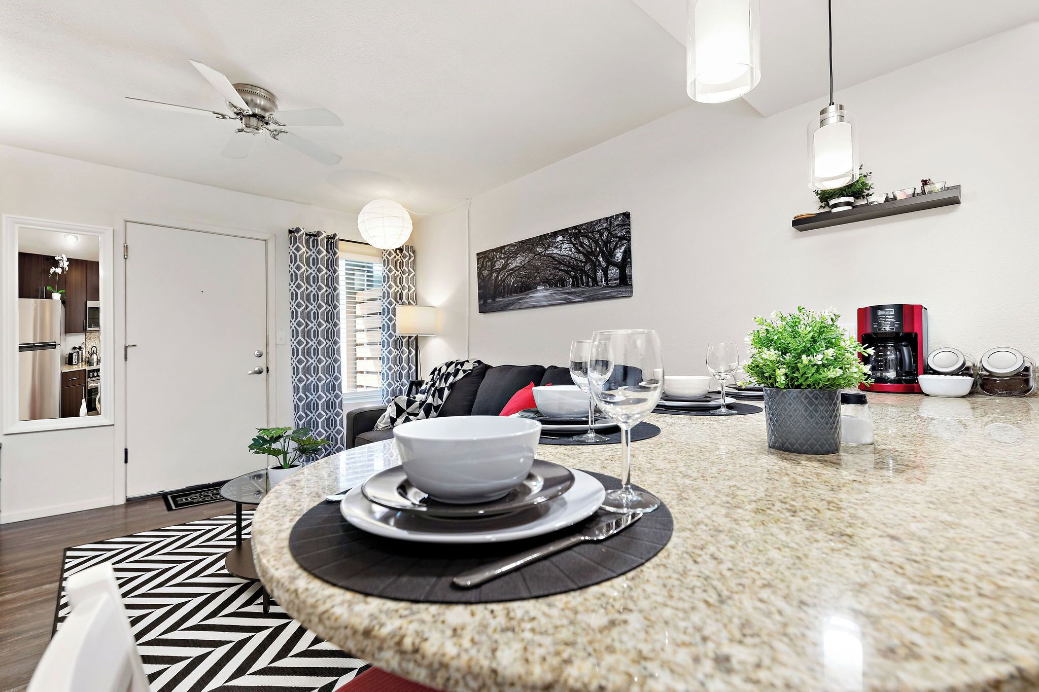 A modern kitchen with a granite countertop, a dining area with a black and white patterned rug, and a living space with a sofa and a coffee machine.