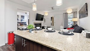Una cocina moderna con encimera de granito, un cubo de basura rojo y una zona de comedor con sofá negro y cortinas estampadas.
