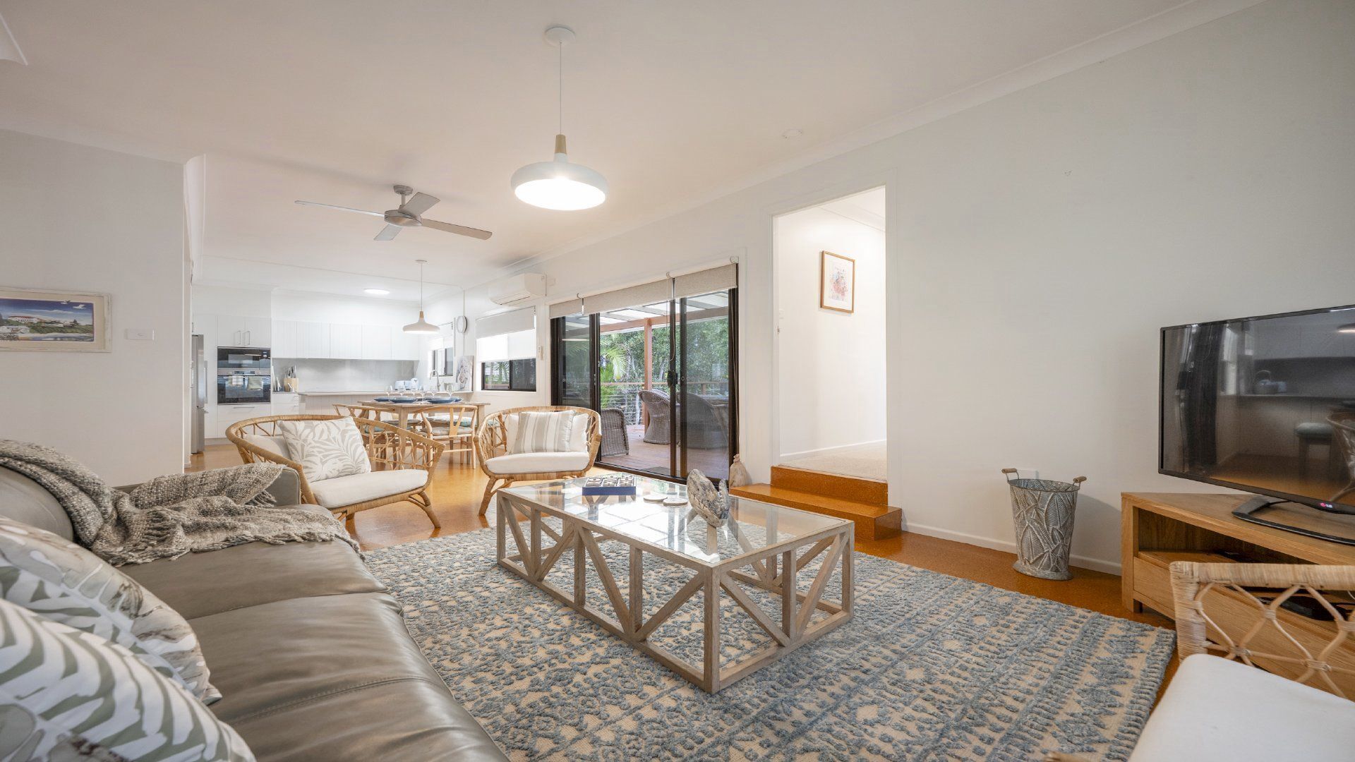 A modern living room with a sofa, a coffee table, and a television. There is a kitchen in the background with white cabinets and a dining area with chairs.