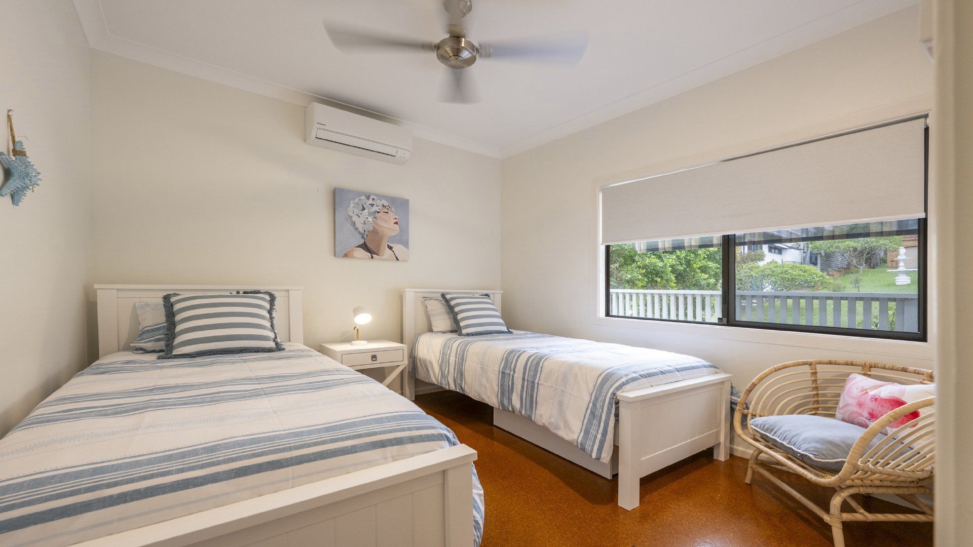 A bedroom with two beds, a ceiling fan, an air conditioning unit, a bedside table with a lamp, a window with a view of greenery, and a wicker chair.