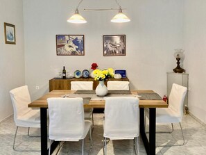 A dining area with a wooden table, white chairs, and a vase of yellow flowers. There are two framed pictures on the wall above the table. - Aphrodite's Apartments (Spata-Artemida)