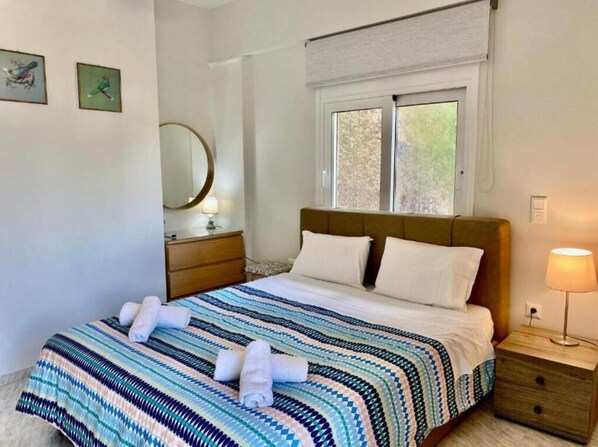 A neatly made bed with a striped bedspread and two pillows, a bedside table with a lamp, and a window with a view of greenery. - Aphrodite's Apartments (Spata-Artemida)