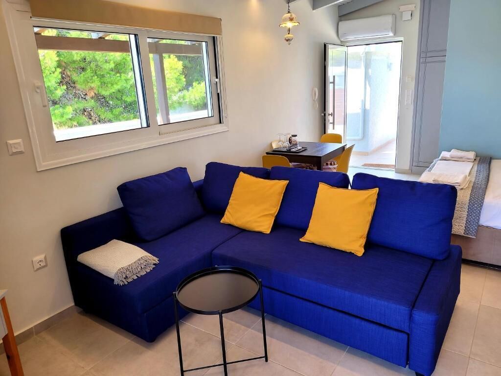 A living room with a blue sofa, yellow cushions, a small round table, and a view of greenery through the windows.