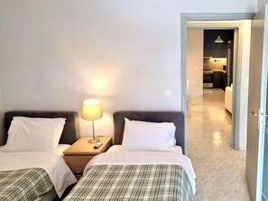 A hotel room with two beds, a nightstand with a lamp, and a view into the kitchen area. - Aphrodite's Apartments (Spata-Artemida)