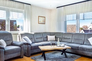 Un salon moderne avec un canapé d’angle gris, une table basse en bois et une vue sur les immeubles à travers de grandes fenêtres.