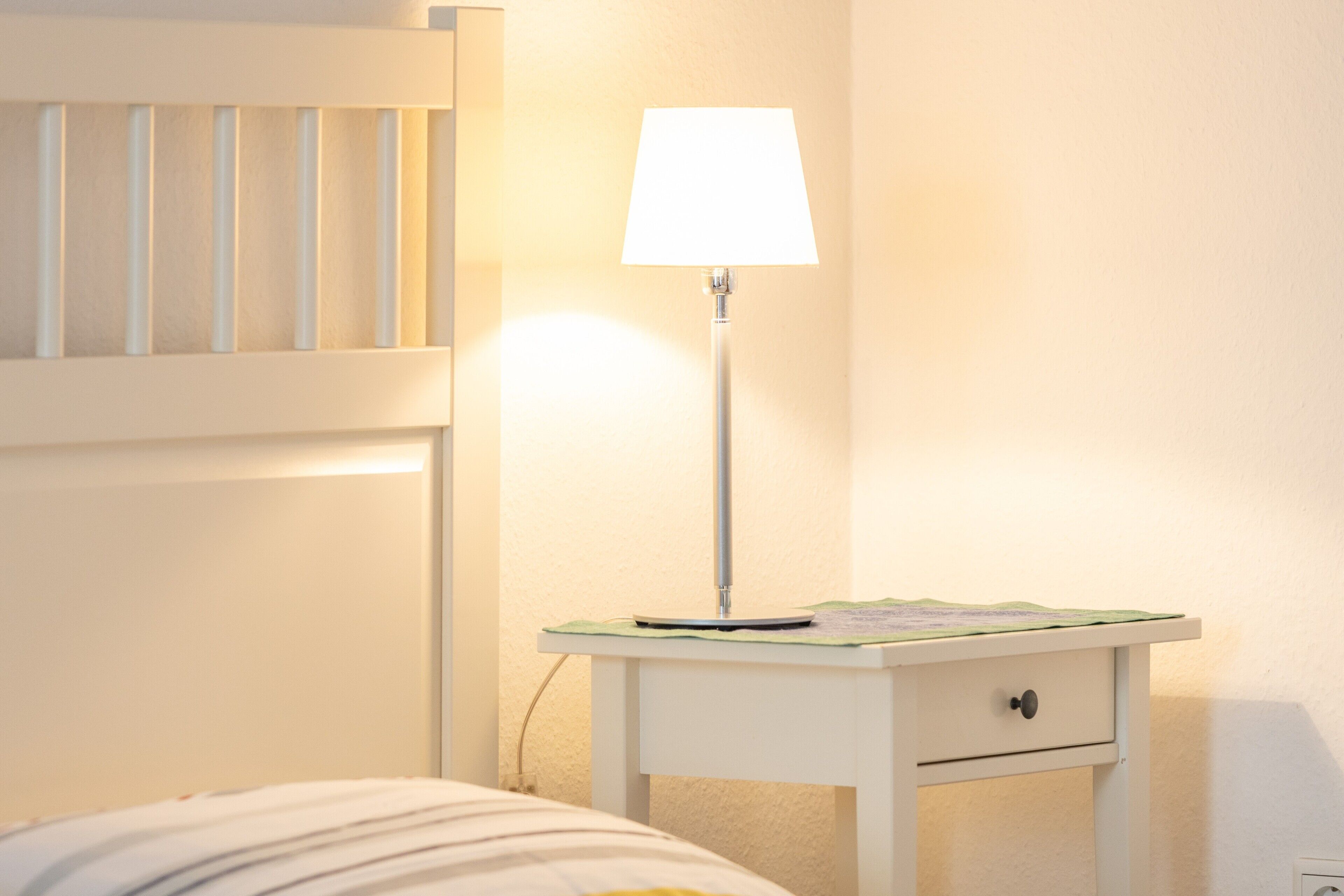 A white bedside table with a lamp, a bed with a striped blanket, and a wall-mounted shelf.