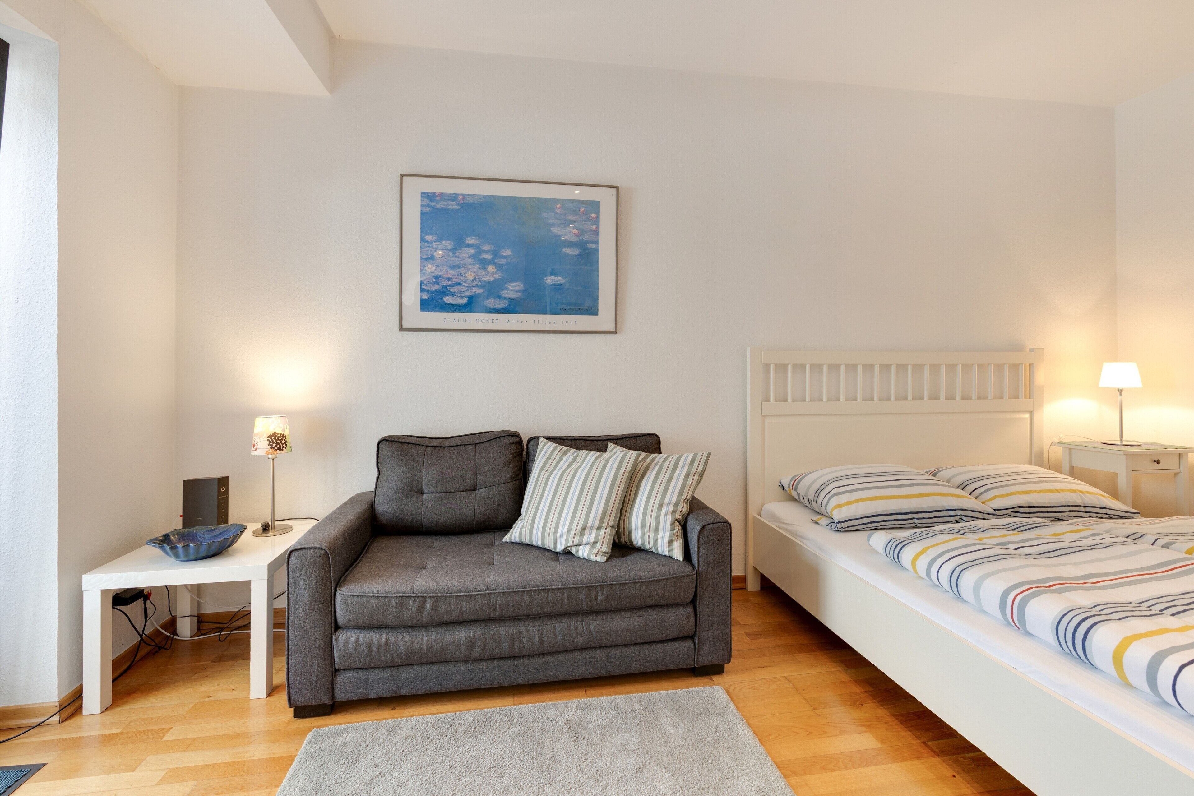 Ein Schlafzimmer mit grauem Sofa, einem Bett mit gestreifter Bettwäsche, einem gerahmten Gemälde über dem Sofa und einem Nachttisch mit Lampe.