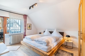 Un dormitorio con cama de madera, ropa de cama blanca, una mesita de noche con lámpara, un radiador y una ventana con cortinas.