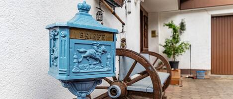 Un buzón azul con la leyenda 'BRIEFE', un carrito de madera y una pared blanca con una puerta al fondo.