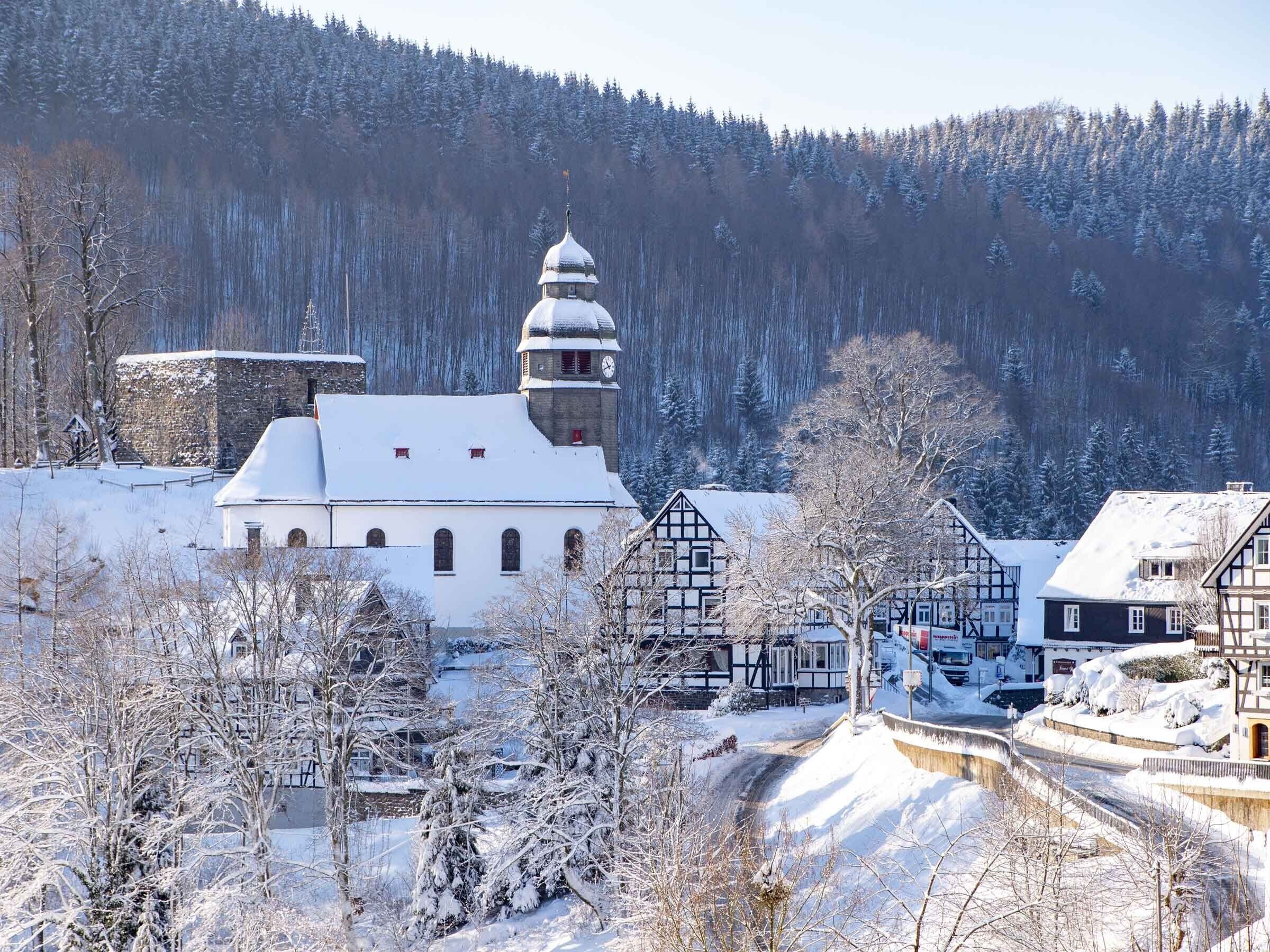 A historic church with a prominent tower, surrounded by traditional timber-framed houses, set in a snowy village.