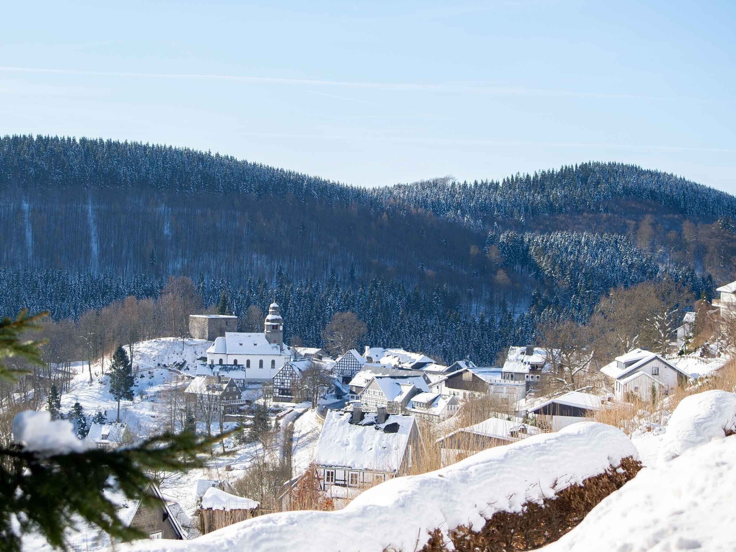 Un villaggio innevato con edifici tradizionali, una chiesa e una collina boscosa sullo sfondo.