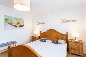 Una cama bien hecha con cabecera de madera, dos mesitas de noche con lámparas, un cuadro de una flor colgado en la pared y la palabra 'Dream' escrita en la pared.