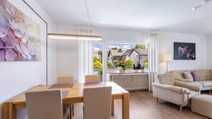 A modern living room with a dining area, a sofa set, and a large window with a view of a house. - Apartment 'Annes Stübchen' with Balcony and Wi-Fi (Schmallenberg)