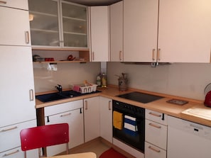 Cocina pequeña con armarios blancos, estufa negra, lavamanos y una silla roja.