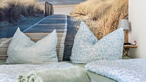 Un lit avec une literie à motifs, des oreillers et une serviette enroulée. Une promenade en bois mène à la plage.