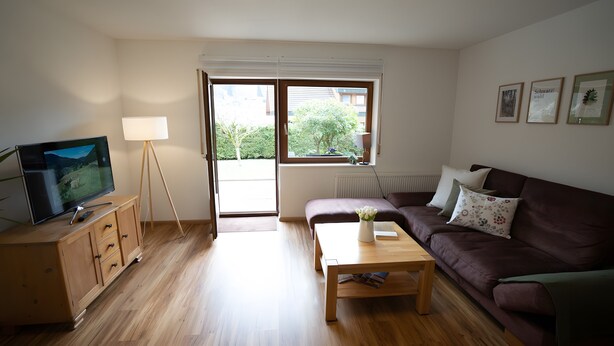 Ein Wohnzimmer mit Fernseher, Stehlampe, Sofa, Couchtisch und einem Fenster mit Blick auf Grünflächen.