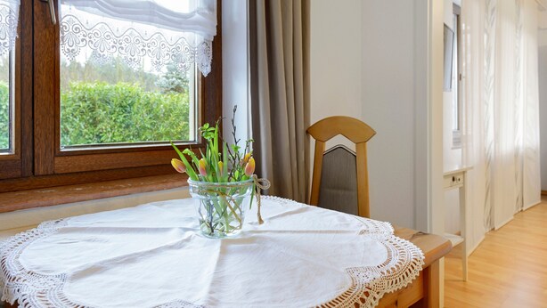 A room with a wooden table, a lace tablecloth, a clear glass vase with flowers, and a view of greenery through a window with curtains. - Apartment 'Thum' with Private Terrace and Wi-Fi (Daisendorf)