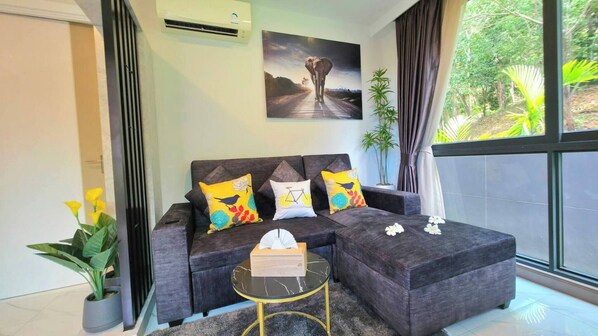A modern living room with a dark grey sectional sofa, colorful cushions, a small round coffee table, and a painting of an elephant on the wall. - Room-a301- 1 Bedroom Corner Condo Forest (Krabi)