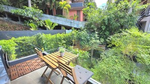A balcony with a wooden table and chairs, surrounded by lush greenery and a view of a building with a red facade. - Room-a301- 1 Bedroom Corner Condo Forest (Krabi)