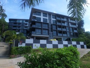 A modern multi-story building with a checkered wall design, surrounded by greenery and palm trees. - Room-a301- 1 Bedroom Corner Condo Forest (Krabi)