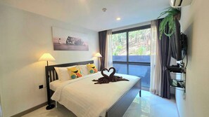 A hotel room with a bed, two bedside lamps, a painting, a TV, and a balcony with curtains. - Room-a301- 1 Bedroom Corner Condo Forest (Krabi)