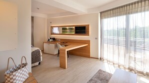 A modern hotel room with a wooden desk, a bed, a TV mounted on the wall, and a large window with curtains.