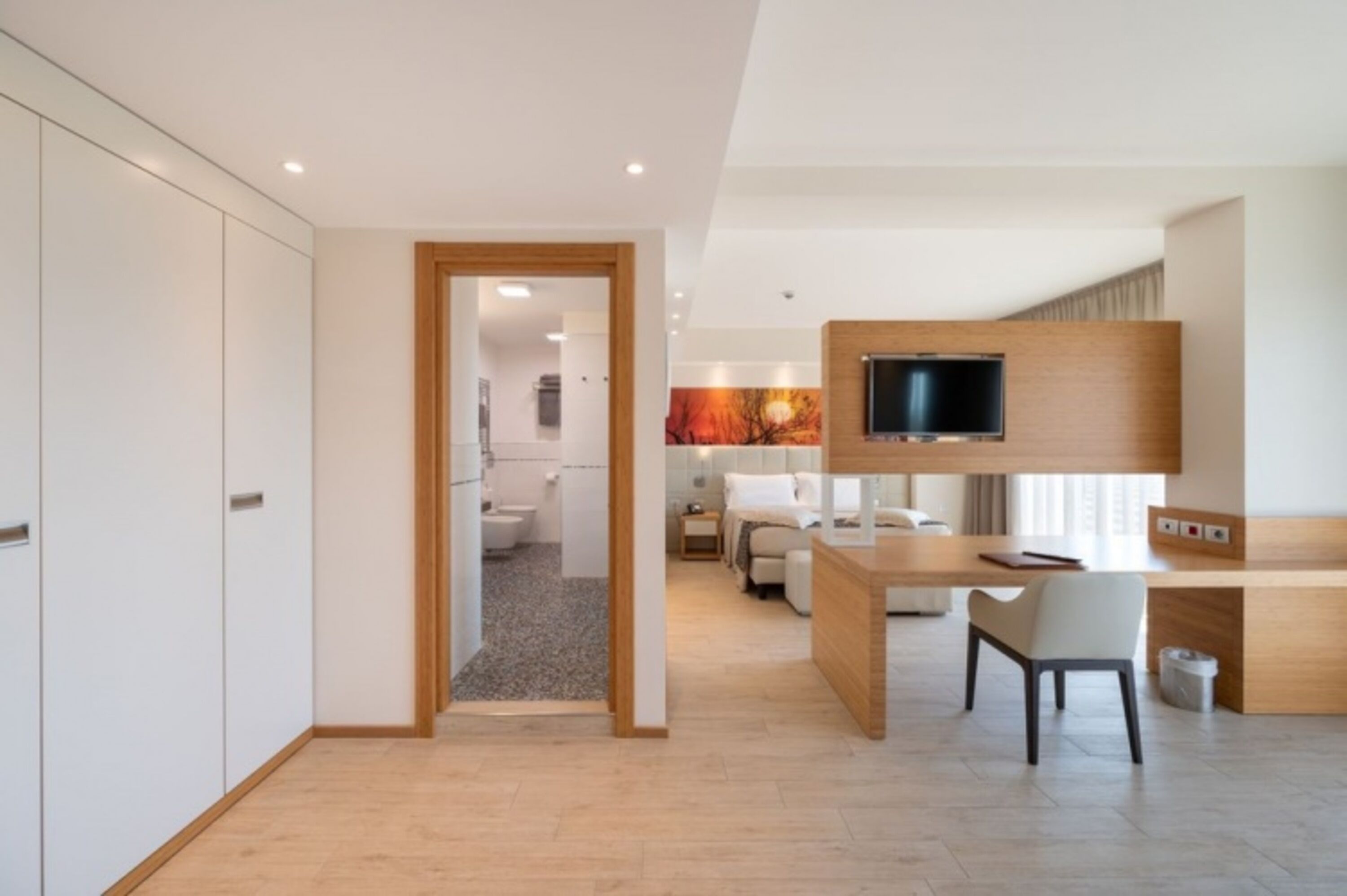 Habitación de hotel moderna con un escritorio de madera, un televisor fijado a la pared y un baño separado visible a través de una puerta abierta.