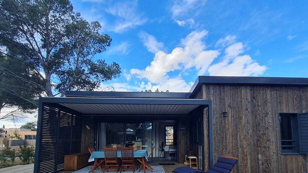 A wooden deck with a dining table and chairs, a covered seating area with lounge chairs, and a view of a modern building with a wooden exterior. - Agde Prestige Chalet in private residence with Pools and Spa (Agde)