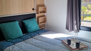A bedroom with a bed, teal pillows, a bedside table with a tray of mugs, a hanging lamp, and a view of greenery outside. - Agde Prestige Chalet in private residence with Pools and Spa (Agde)