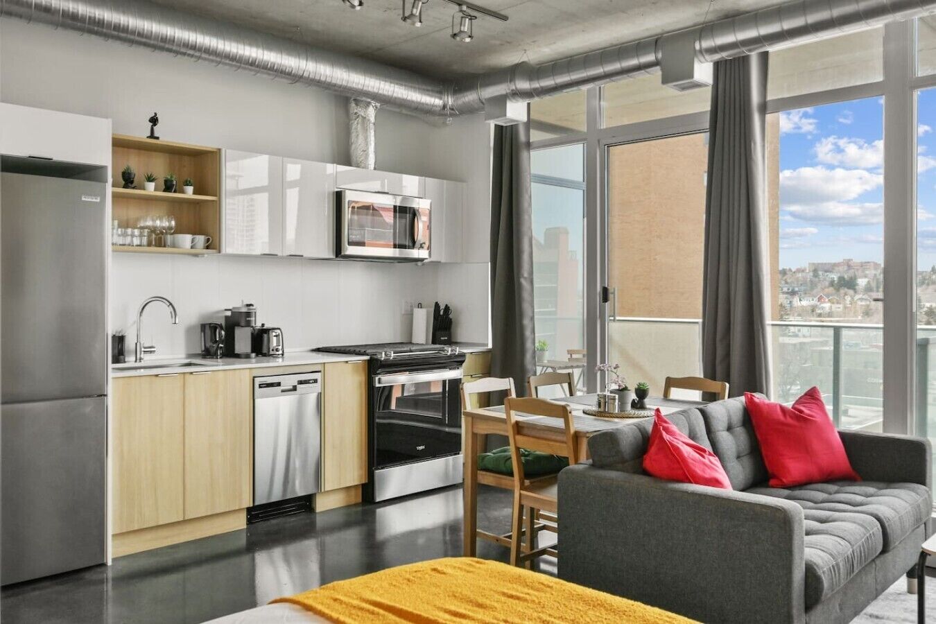 A modern kitchen with a microwave, oven, and dishwasher, a dining area with a table and chairs, and a gray sofa with red cushions.