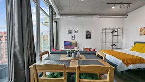 A modern hotel room with a dining table, chairs, a bed, a TV, artwork on the wall, and a view of the city. - Bright Studio | Mins to Saddledome | Skyline Views (Calgary)