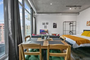 A modern hotel room with a dining table, chairs, a bed, a TV, artwork on the wall, and a view of the city. - Bright Studio | Mins to Saddledome | Skyline Views (Calgary)