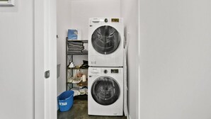 A laundry room with two stacked washers and dryers, a blue bucket, and a shelf with folded clothes. - Bright Studio | Mins to Saddledome | Skyline Views (Calgary)