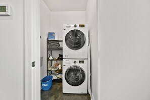 A laundry room with two stacked washers and dryers, a blue bucket, and a shelf with folded clothes. - Bright Studio | Mins to Saddledome | Skyline Views (Calgary)