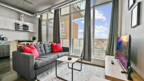 A modern living room with a grey sofa, red cushions, a small table, and a TV on a stand. - Bright Studio | Mins to Saddledome | Skyline Views (Calgary)
