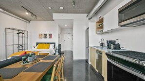 A modern kitchen with a built-in stove, a dining area with a wooden table and chairs, and a bed with a yellow pillow. - Bright Studio | Mins to Saddledome | Skyline Views (Calgary)